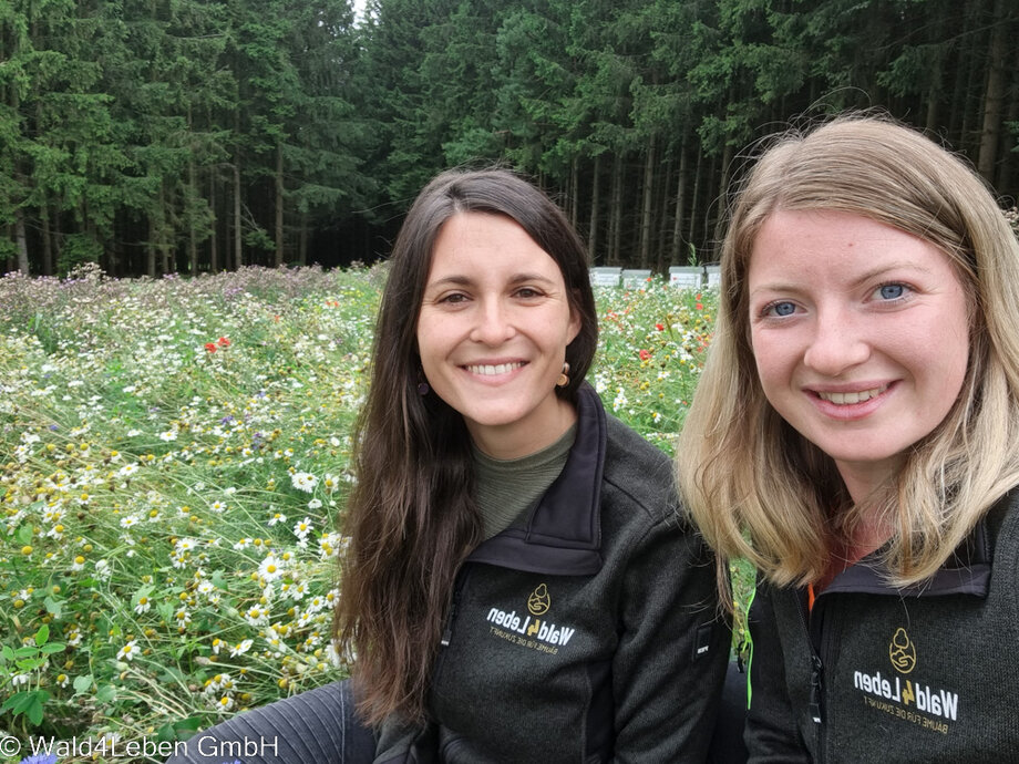 Wald4Leben Mitarbeiterinnen Nadja und Magdalena auf der Blühfläche in Götzles mit den Bienenstöcken im Hintergrund.